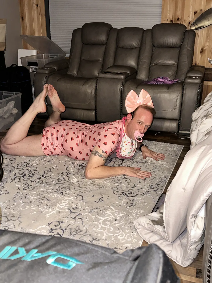 A person in pink lies on a patterned rug in front of a reclining chair in a cluttered room.