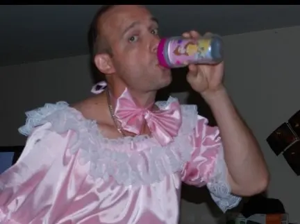A man in a pink satin frilly outfit drinks from a bottle in a dim room.