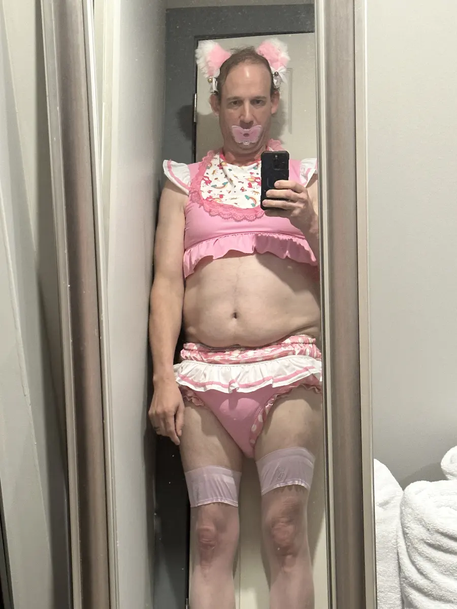 A person takes a mirror selfie in a pink frilly outfit with matching cat-ear accessories and a pink pacifier-style mouthpiece.