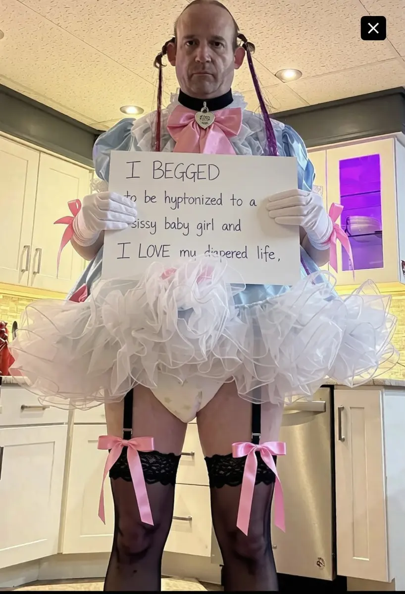 A person in a ruffled costume and gloves holds a handwritten sign in a bright kitchen.