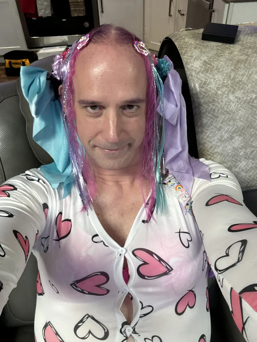 A close selfie shows a person with pink and purple hair accessories wearing a white heart-print top.