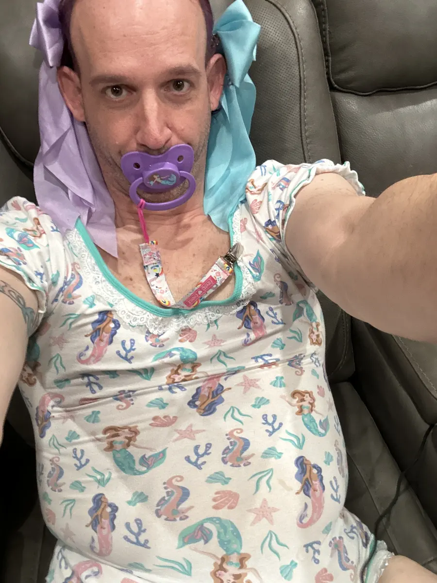 A selfie shows a person on a leather seat wearing a mermaid-print outfit and a purple pacifier.