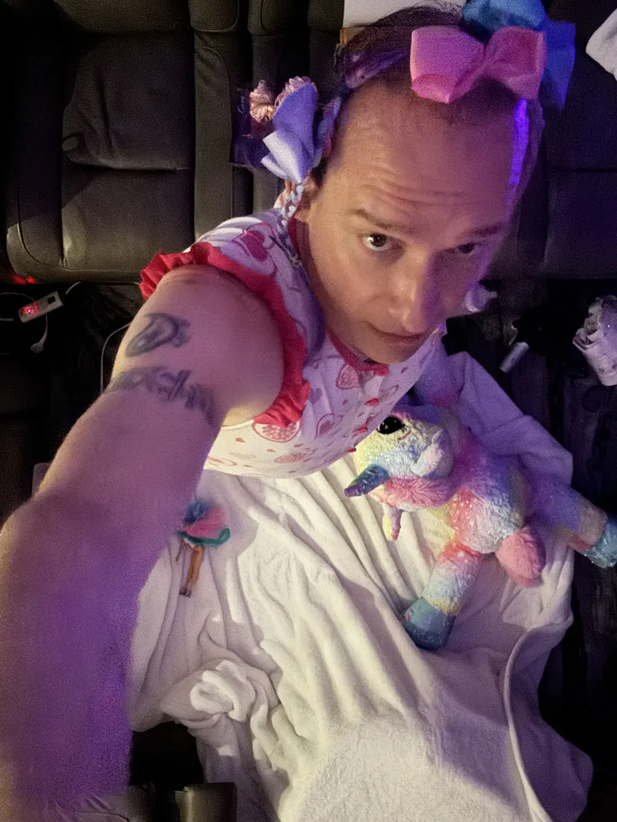 An overhead selfie shows a person in a heart-print outfit beside a pastel unicorn plush inside a dim vehicle.
