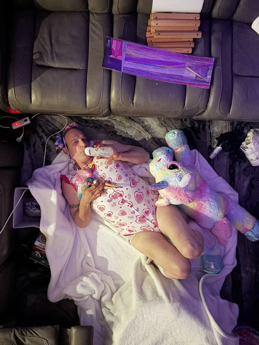 A person lies on a white blanket in a vehicle interior with a bottle, toy, and pastel unicorn plush nearby.