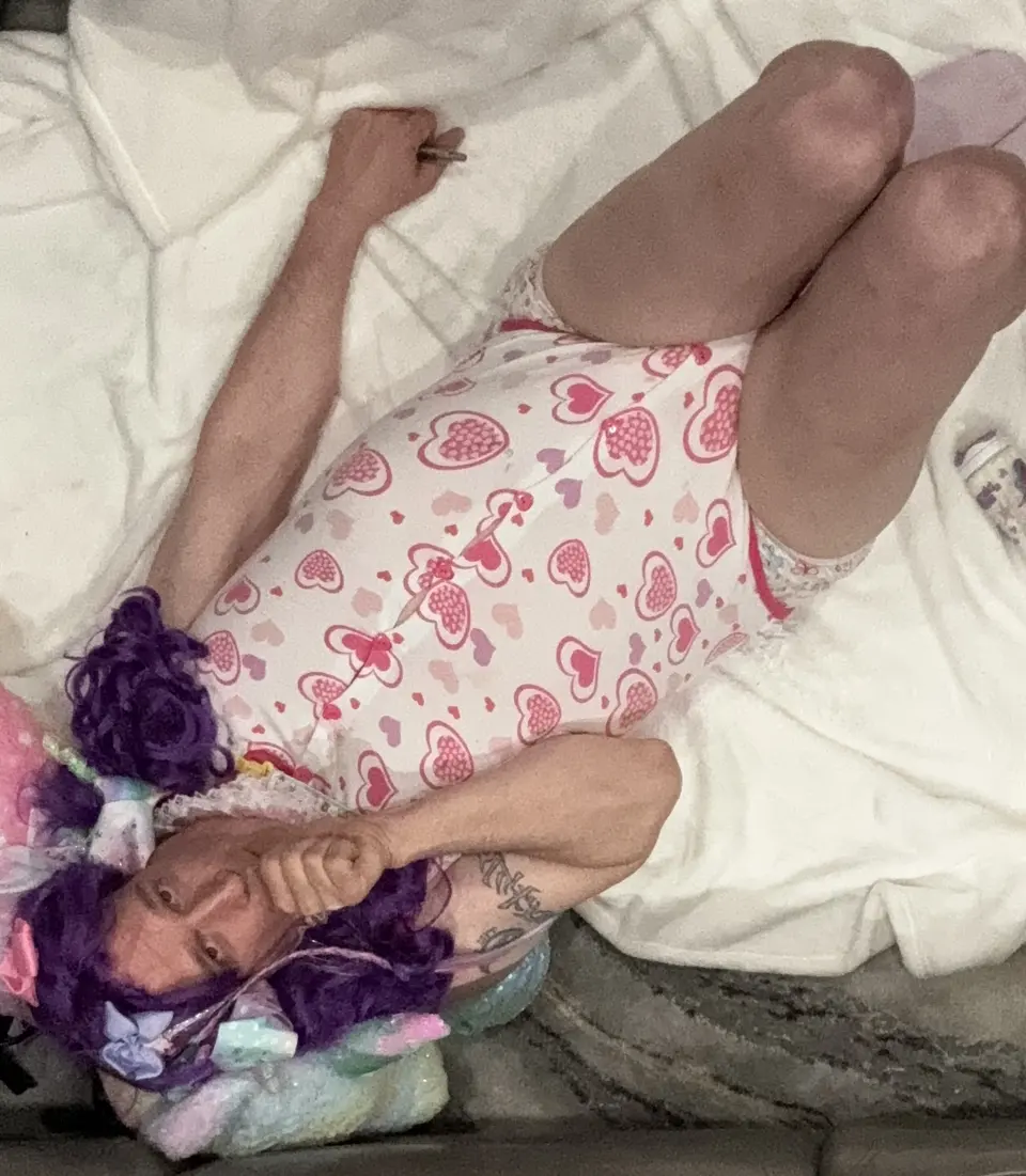 A person reclines on white bedding in a pink heart-print outfit, with purple hair accessories and a bent arm near the face.