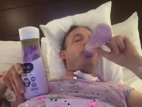 A person reclines on white pillows, holding a lavender bottle and a bottle-shaped object near the mouth.