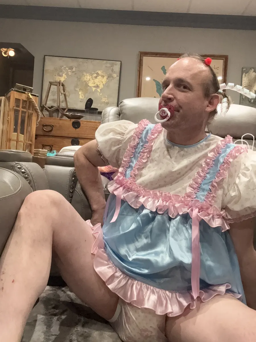 A man in a pastel ruffled outfit reclines indoors with a pacifier and one knee raised.