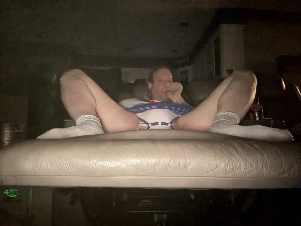 A person in a sailor-style outfit lounges on a leather couch in a dark room, feet in white socks near the camera.