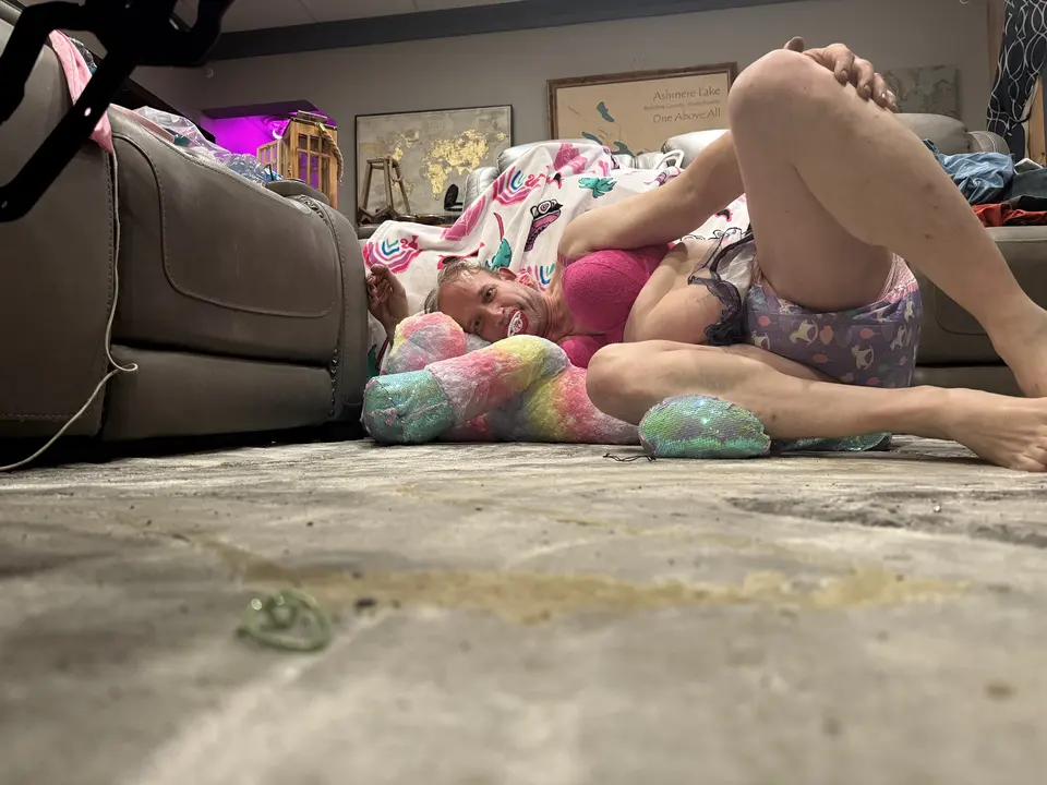 A person wearing a pink top lies on the floor with a pacifier, framed by a sofa and pastel plush toy.