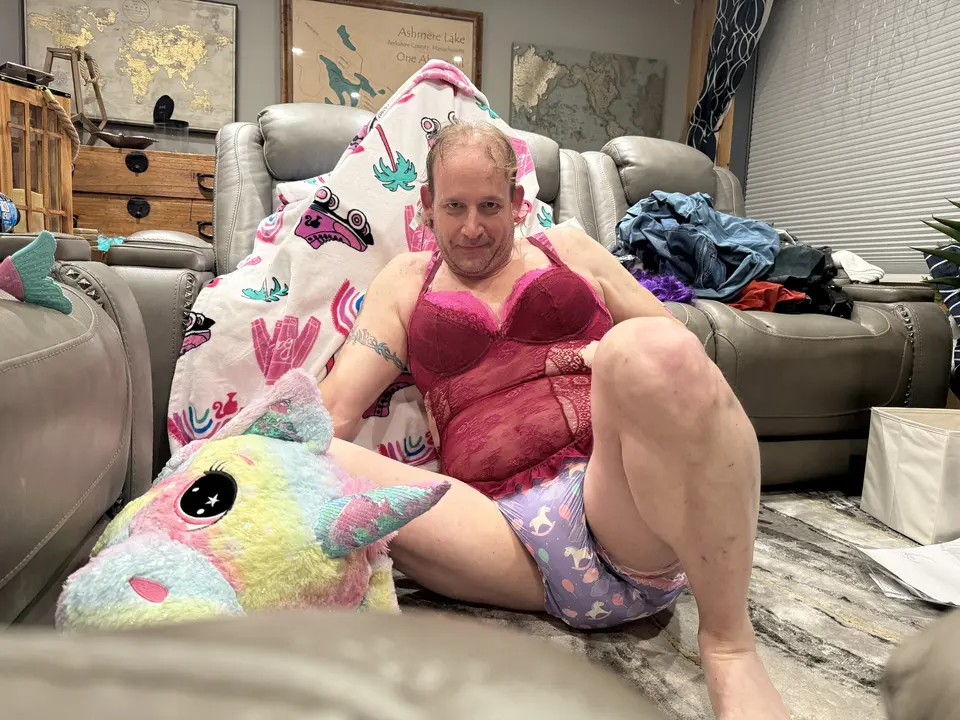 A person in a magenta lace top reclines on the floor beside a rainbow plush toy and a recliner.