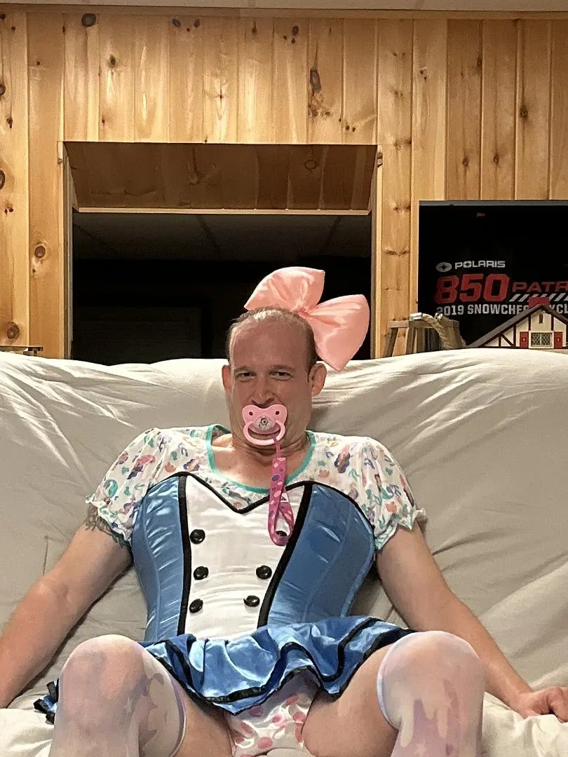 Person seated in a blue costume dress, pink bow, and pacifier in a wood-paneled room.