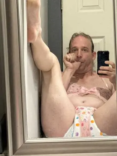 Mirror selfie of a person posing with one leg raised, wearing pastel underwear and a pink bra.