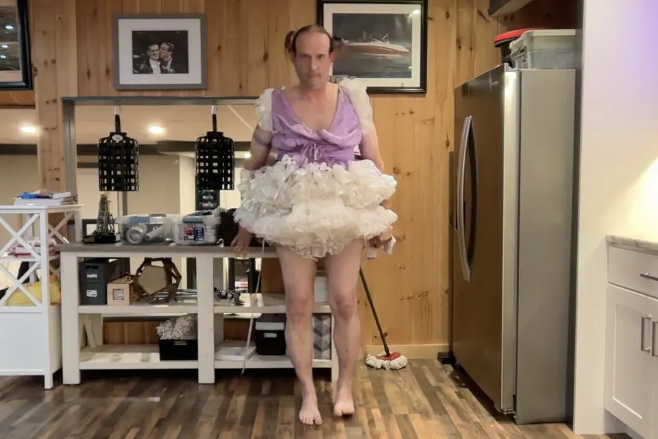 A barefoot person in a lavender top and white ruffled skirt stands in a kitchen with wood-paneled walls.