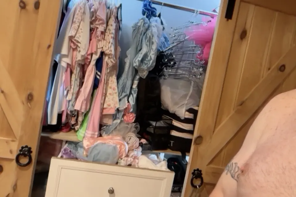 An open closet displays pastel clothing, satin garments, and packed hangers beside a wood-paneled doorway.