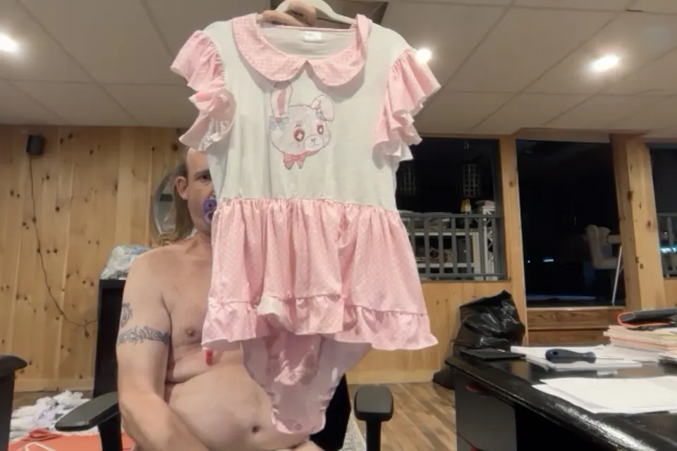 A shirtless person holds up a pink-and-white ruffled dress with a cartoon animal print indoors.