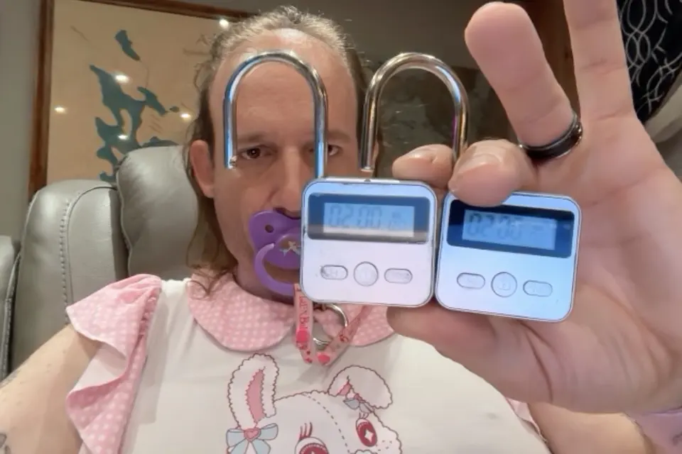A seated person holds two small digital padlocks toward the camera while wearing a purple pacifier.