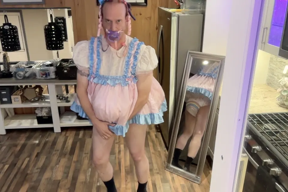 A person in a pink-and-blue frilly outfit stands in a room beside a full-length mirror that reflects the back of the outfit.