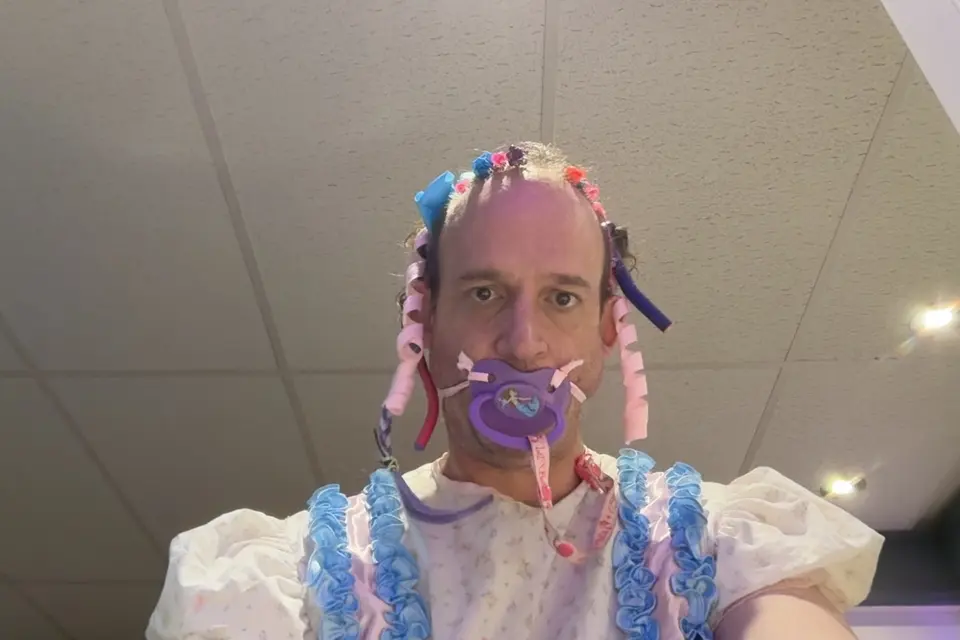 A close-up portrait shows a person with a frilly headpiece and a purple pacifier against a ceiling background.
