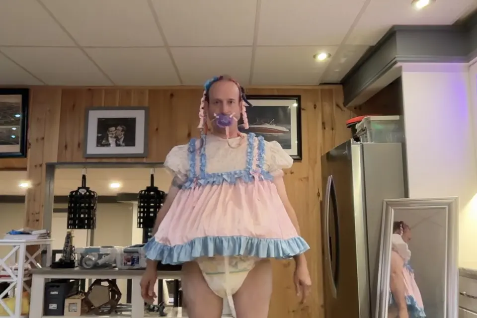 A person stands in a kitchen wearing a loose pink-and-blue frilled dress with a visible diaper underneath.