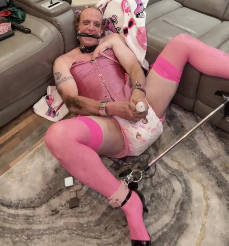 A person reclines on a rug in pink stockings and a corset top, holding a white bottle-shaped object.