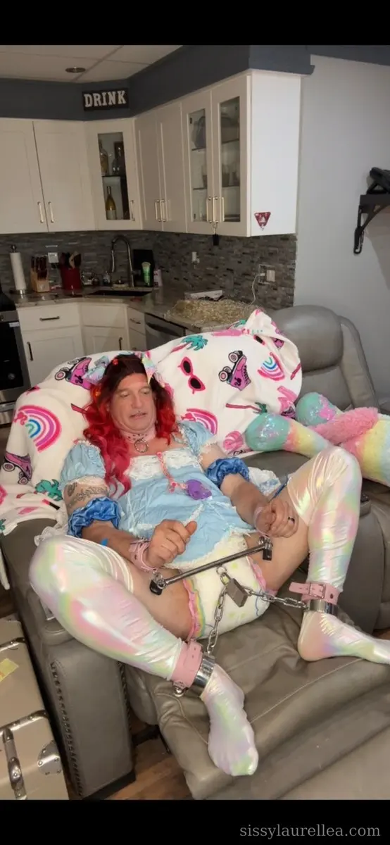 Person lounging in a gray recliner wearing a pastel blue dress, shiny leggings, and pink accessories in a room with white cabinets.