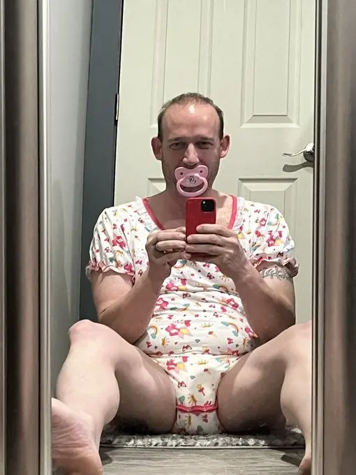 Seated mirror selfie of a person with a pink pacifier, a white printed outfit, and a red phone in front of a paneled door.